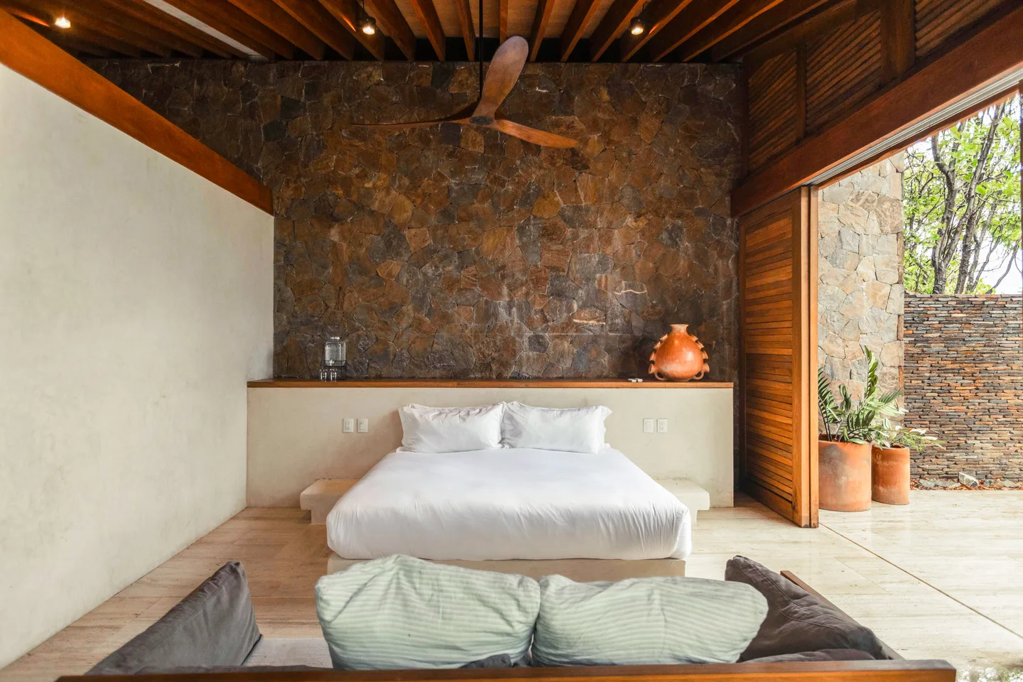King bed beneath a stone wall in Suite Olas, with lounge cushions in the foreground — La Valise Mazunte