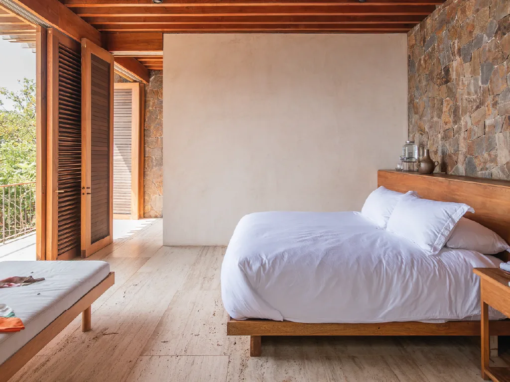 King bed with white linen against a stone wall, louvred wooden doors open to the terrace and a daybed in the foreground in Suite Persianas — La Valise Mazunte
